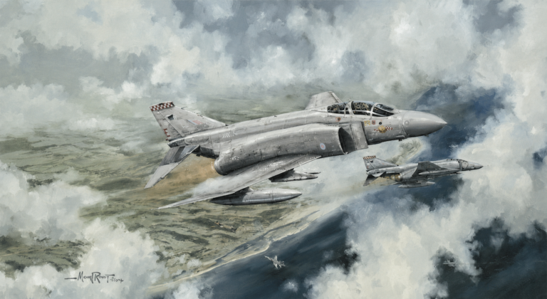 Twin-seat gray military jet flying through cloudy sky above coastline, with rear aircraft in formation and drop tanks under wings