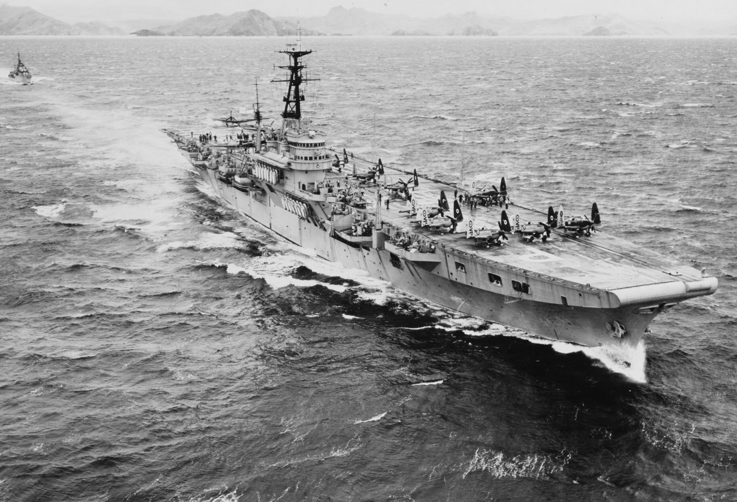 Warship at sea with planes parked on its flight deck and small boats nearby, in a calm, distant island-scape.