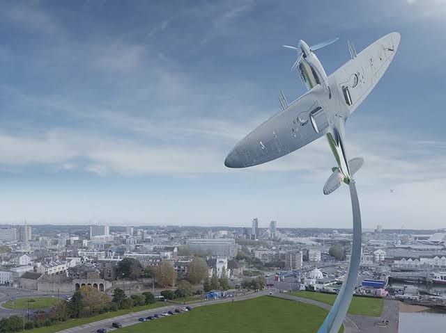 Giant metallic airplane sculpture arcing over a cityscape with roads and buildings below.