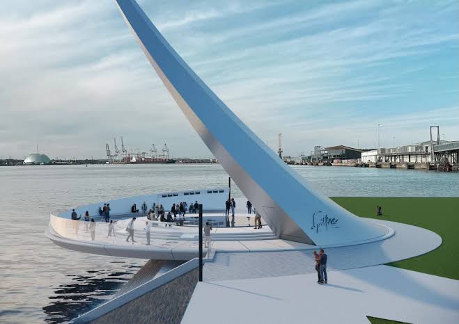 Public waterfront plaza with a tall blue diagonal arch sculpture over a curved white platform and people walking nearby by the harbor.